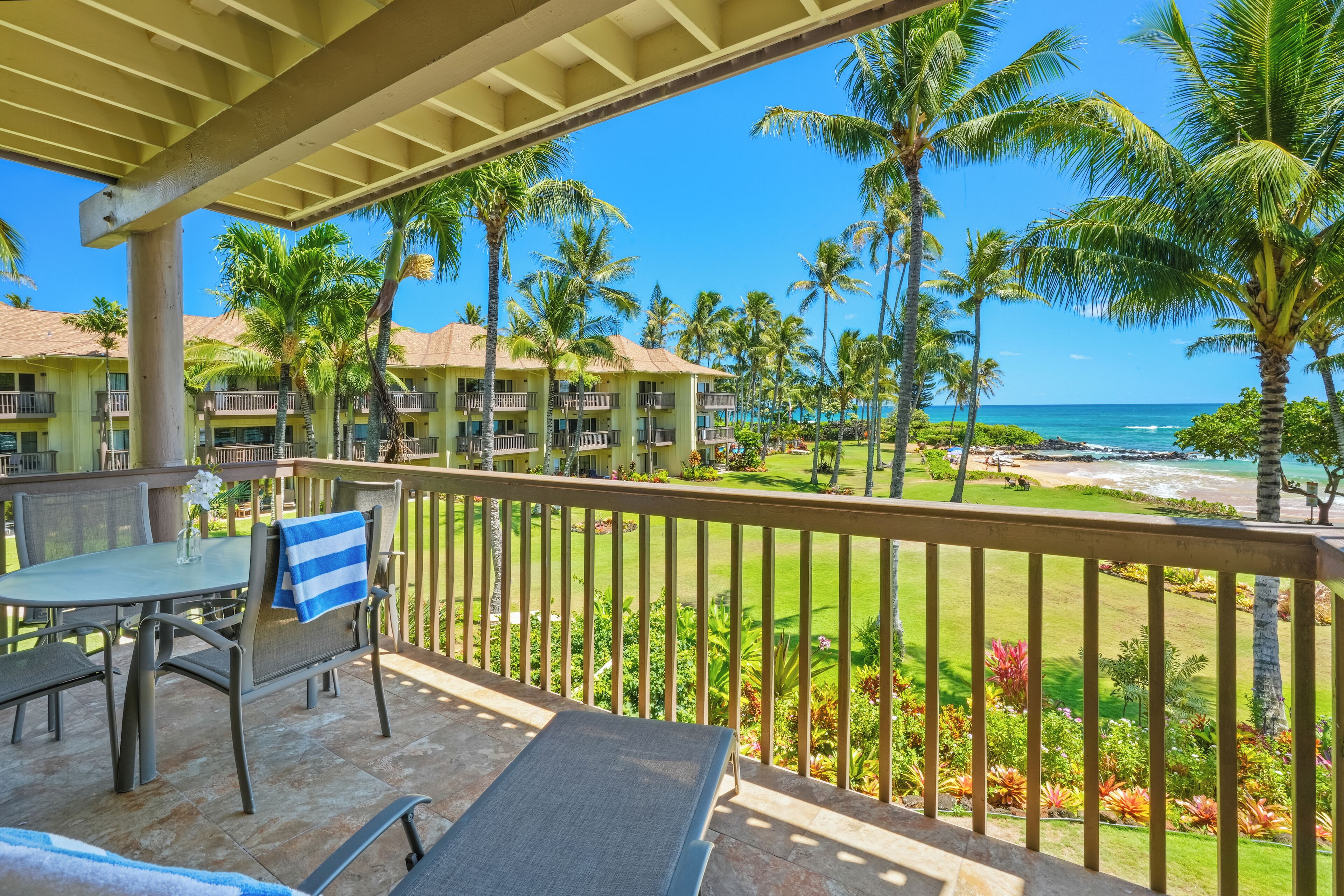 A balcony with a table and chairs, overlooking a beach with palm trees and a clear blue sky.