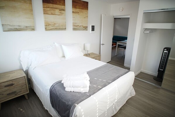 A neatly made bed with white linens and a gray blanket, a bedside table with a lamp, and a wooden nightstand.