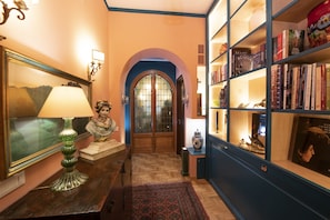 Un couloir doté d’un plancher en bois, une statue posée sur une table, une bibliothèque et une porte ornée de vitraux.