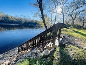 Une terrasse en bois avec une rambarde en métal, donnant sur un lac paisible, entouré d’arbres et de zones herbeuses.