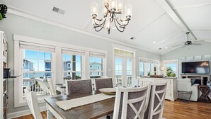 A spacious kitchen with a large island, a dining area with a wooden table and chairs, and a living area with a sofa and TV. - Pier Walker: Spacious, Beautiful Home with Amazing Ocean Views Steps from OKI Pi (Oak Island)