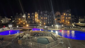 A nighttime view of a resort with two large swimming pools, a covered deck area, and a series of multi-story buildings with balconies. - BIS-Duck Resort - Spacious 2BR/2BA with Pool, Courtyard, Ocean View (Duck)