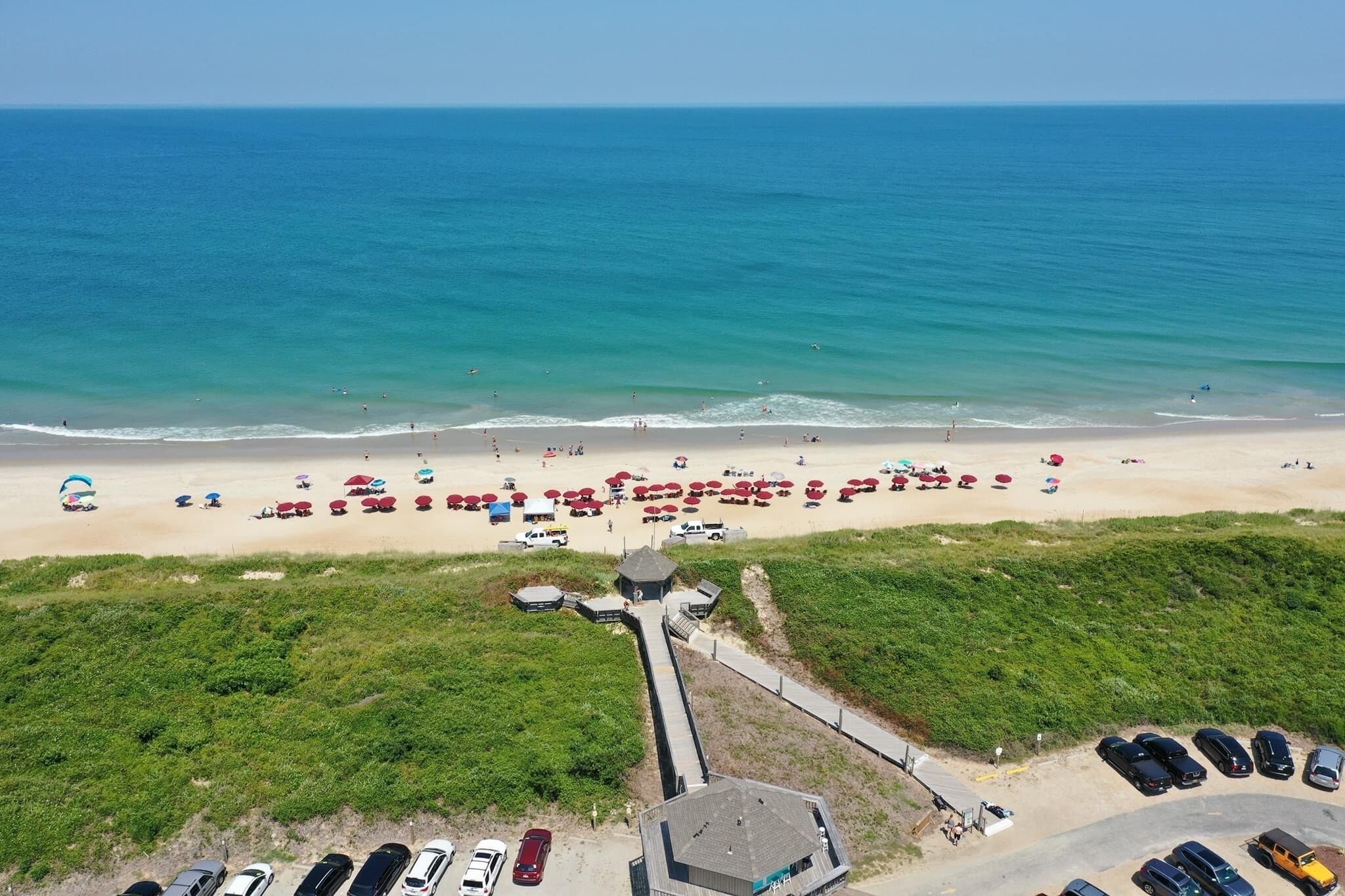 Vista aérea de una playa con hileras de sombrillas y sillas de playa, un estacionamiento y un pequeño edificio cerca de la orilla.