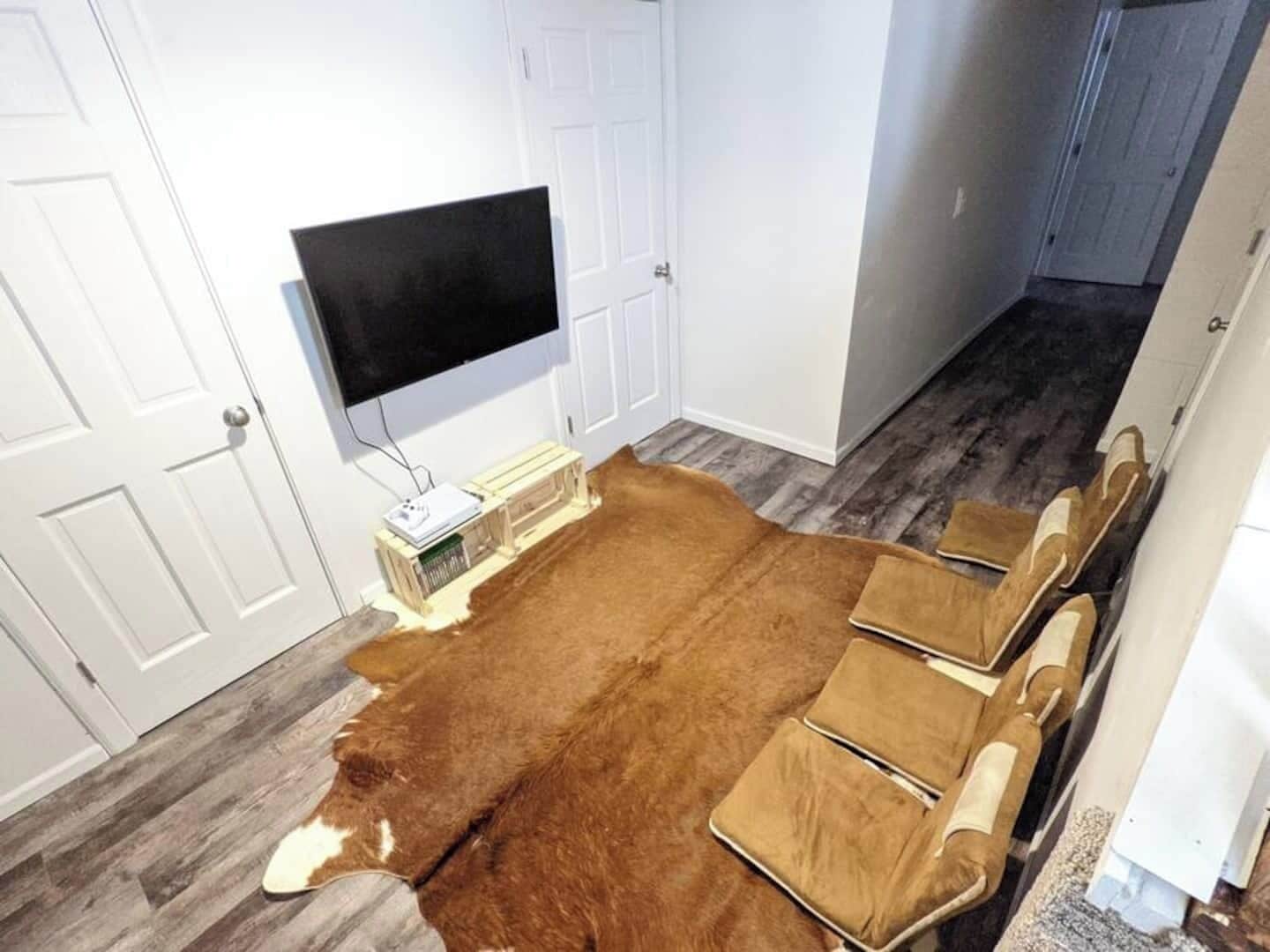 A room with a large brown rug, white walls, a mounted TV, and a wooden bench with cushions.