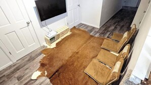 Una habitación con un tapete marrón grande, paredes blancas, un televisor montado y un banco de madera con cojines.