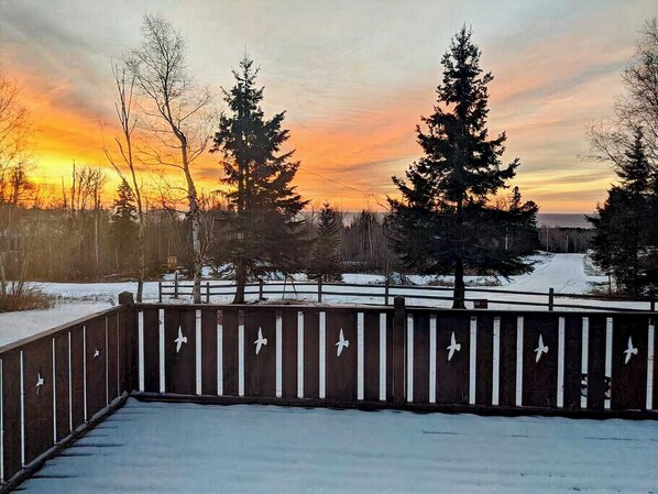 A wooden deck with a railing, overlooking a snowy landscape with trees and a sunset.