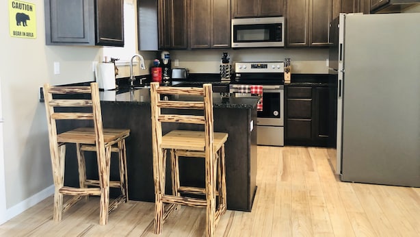 A modern kitchen with dark wood cabinets, a stainless steel refrigerator, a microwave, and a built-in oven. There are two wooden bar stools and a small island with a red and white checkered towel. - Riverside Cabin 6 (Grants Pass)