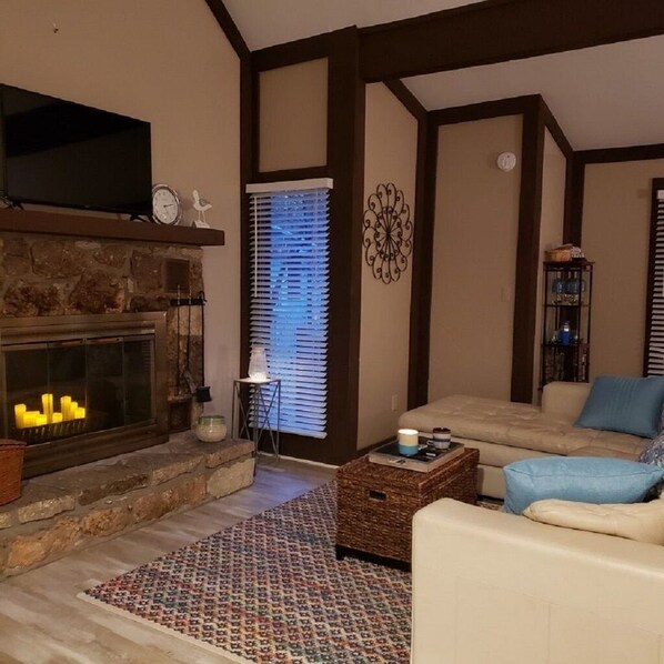 A living room with a stone fireplace, a television mounted above it, a sofa with cushions, a patterned rug, and a decorative wall piece.