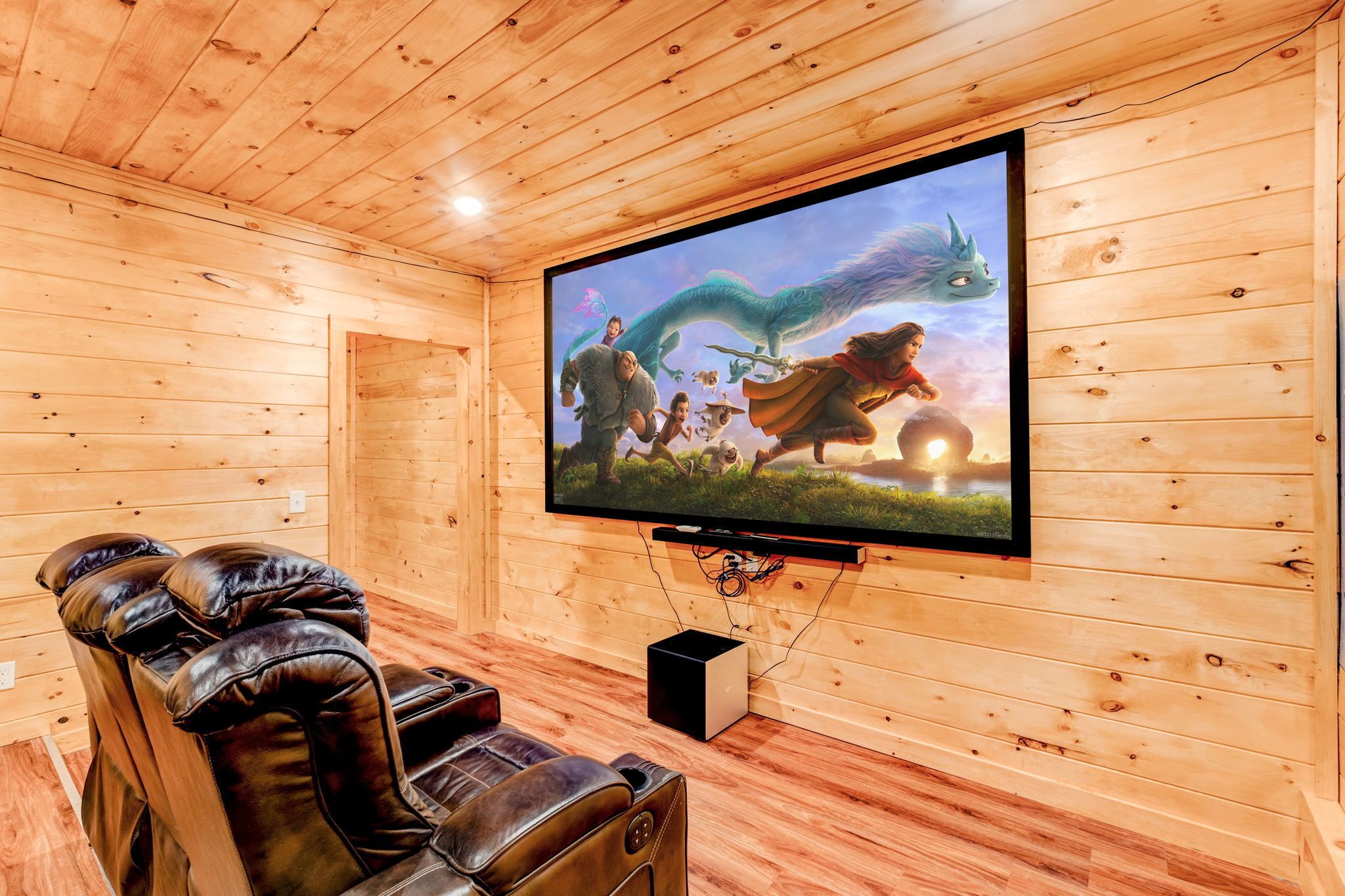 Una habitación con paneles de madera, un televisor de pantalla plana grande montado en la pared, sillones de cuero reclinables y un altavoz negro pequeño.
