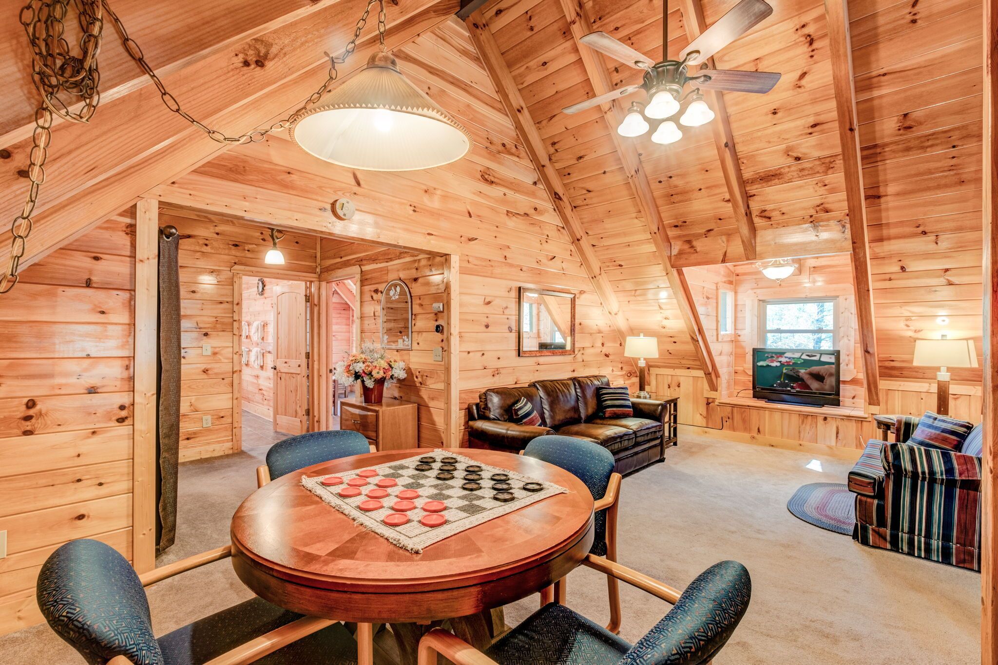 A cozy wooden cabin interior with a dining table, chairs, a leather sofa, a television, and a ceiling fan.