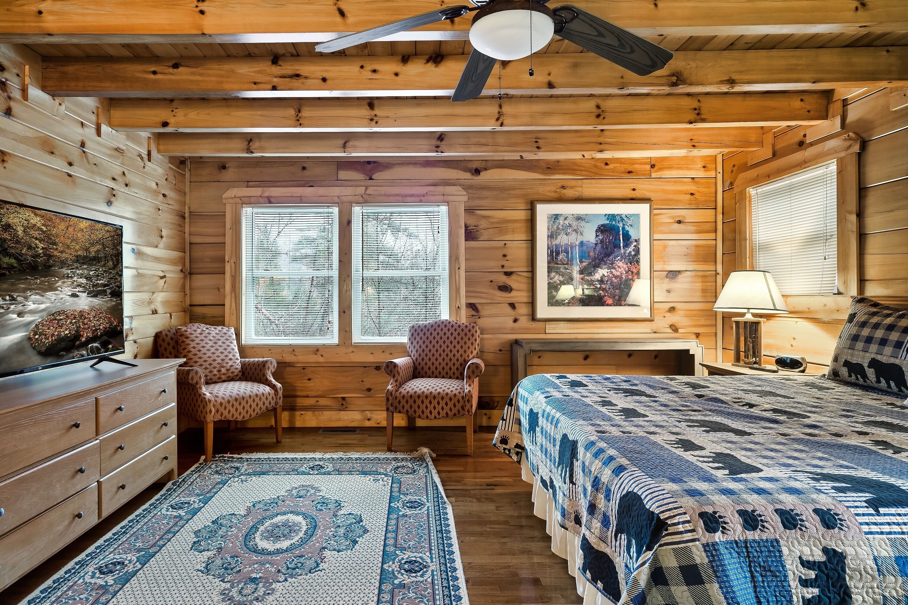A cozy cabin room with wooden walls, a ceiling fan, a bed with a patterned quilt, a chair, a desk with a lamp, and a large window with blinds.