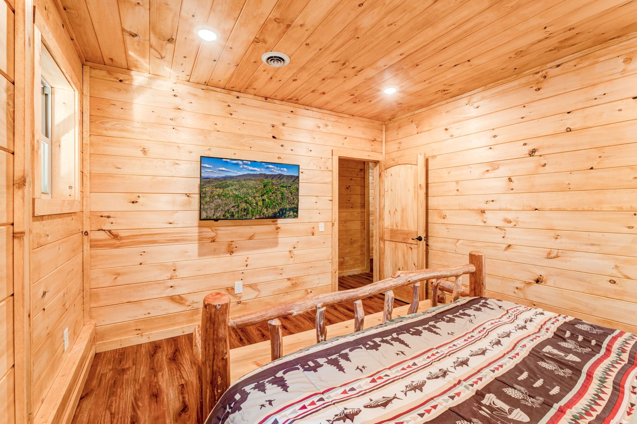A wooden cabin room with a bed, a bench, and a mounted TV displaying a landscape.