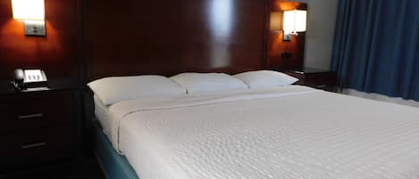 A hotel room with a large bed, wooden headboard, two bedside lamps, a telephone, and a window with blue curtains.