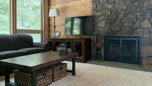 A cozy living room with a stone fireplace, a wooden ceiling, and a large window offering a view of the outdoors. - Good Vibes All Around: Aspen Sixteen at Black Butte Ranch (Black Butte Ranch)