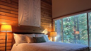 A cozy bedroom with a wooden headboard, a large window offering a forest view, and a wall-mounted sign with positive affirmations. - Good Vibes All Around: Aspen Sixteen at Black Butte Ranch (Black Butte Ranch)