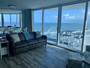 A living room with a large window overlooking the ocean, a dark leather sofa, and a dining area with white chairs and a table.