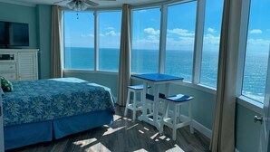A bedroom with a bed, a TV, a ceiling fan, a small table, and chairs. It has large windows with a view of the ocean. - Oceanfront 3-bedroom condo – 17SE - Beach Access, Pool & Hot tub! Owner Managed (Daytona Beach Shores)