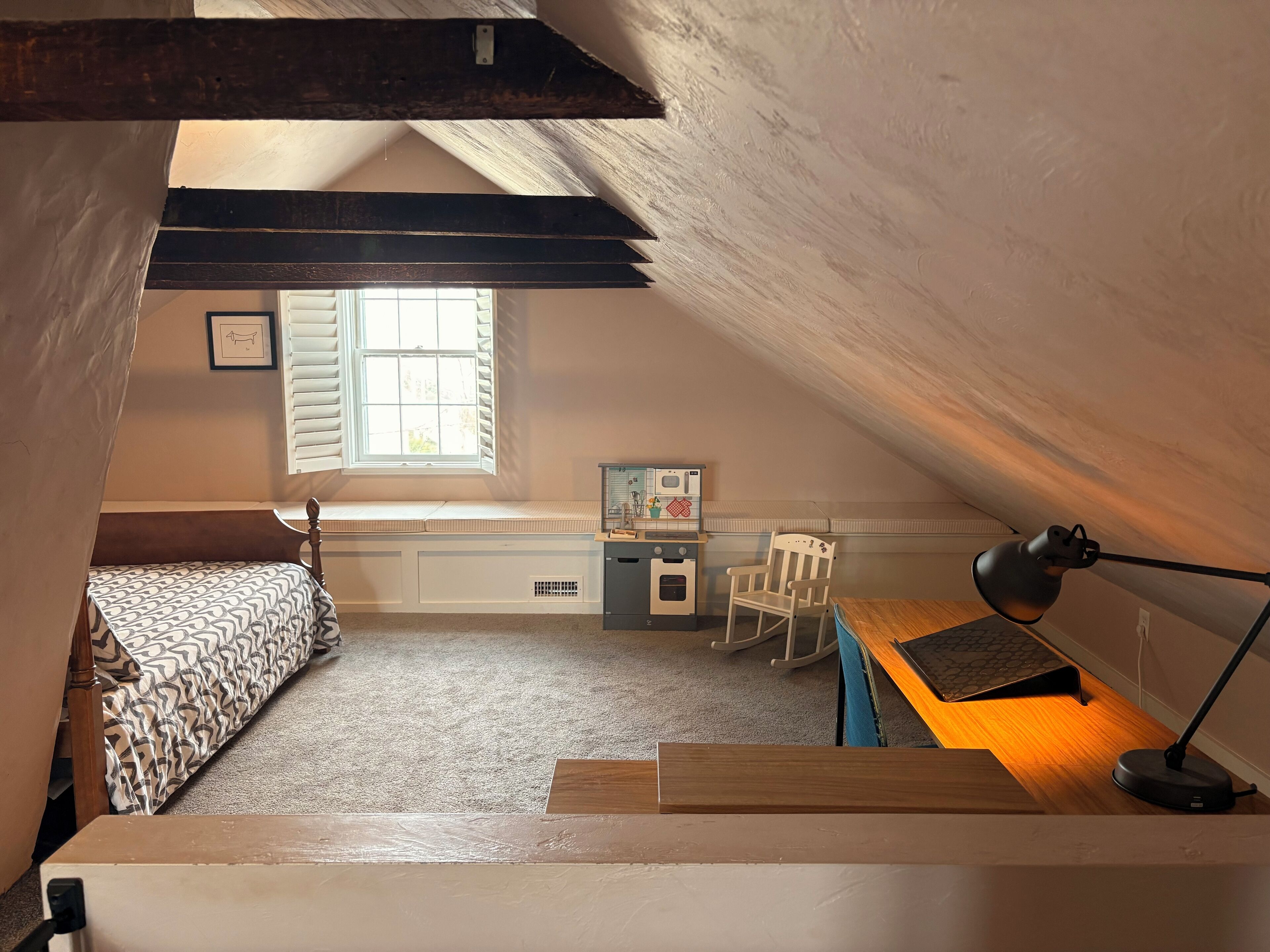 A cozy attic bedroom with a sloped ceiling, a window with shutters, a bed with patterned bedding, a desk with a lamp, and a rocking chair.