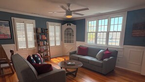 A living room with a gray sofa, a coffee table, and a bookshelf. There are two windows with blinds, a ceiling fan, and a door with a glass panel.