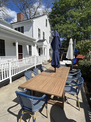 Een houten terras met een tafel en blauwe stoelen, een wit hek en een huis op de achtergrond.