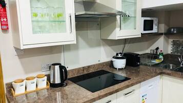 Una cocina con una placa de inducción negra, un microondas, un hervidor de agua y una cafetera. Hay armarios con puertas de vidrio y un lavamanos.