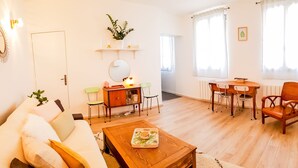 A well-lit living room with a sofa, coffee table, dining table, and chairs. There is a small plant on a shelf, a mirror above the sofa, and a radiator near the windows. - Cosy apartment for 1 to 4 people near the CHU (Rouen)