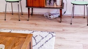 A room with a wooden coffee table, two green chairs, a white rug, a wooden cabinet with glass doors, a round mirror, and a potted plant on a white shelf. - Cosy apartment for 1 to 4 people near the CHU (Rouen)