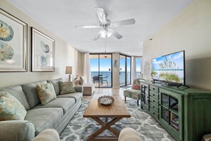 Ein Wohnzimmer mit Sofa, Couchtisch und Fernseher. An den Wänden hängen Bilder in Rahmen und es gibt einen Deckenventilator.