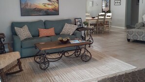 Una sala de estar con un sofá azul, una mesa de centro de madera, un ventilador de techo y una zona de comedor con mesa y sillas.