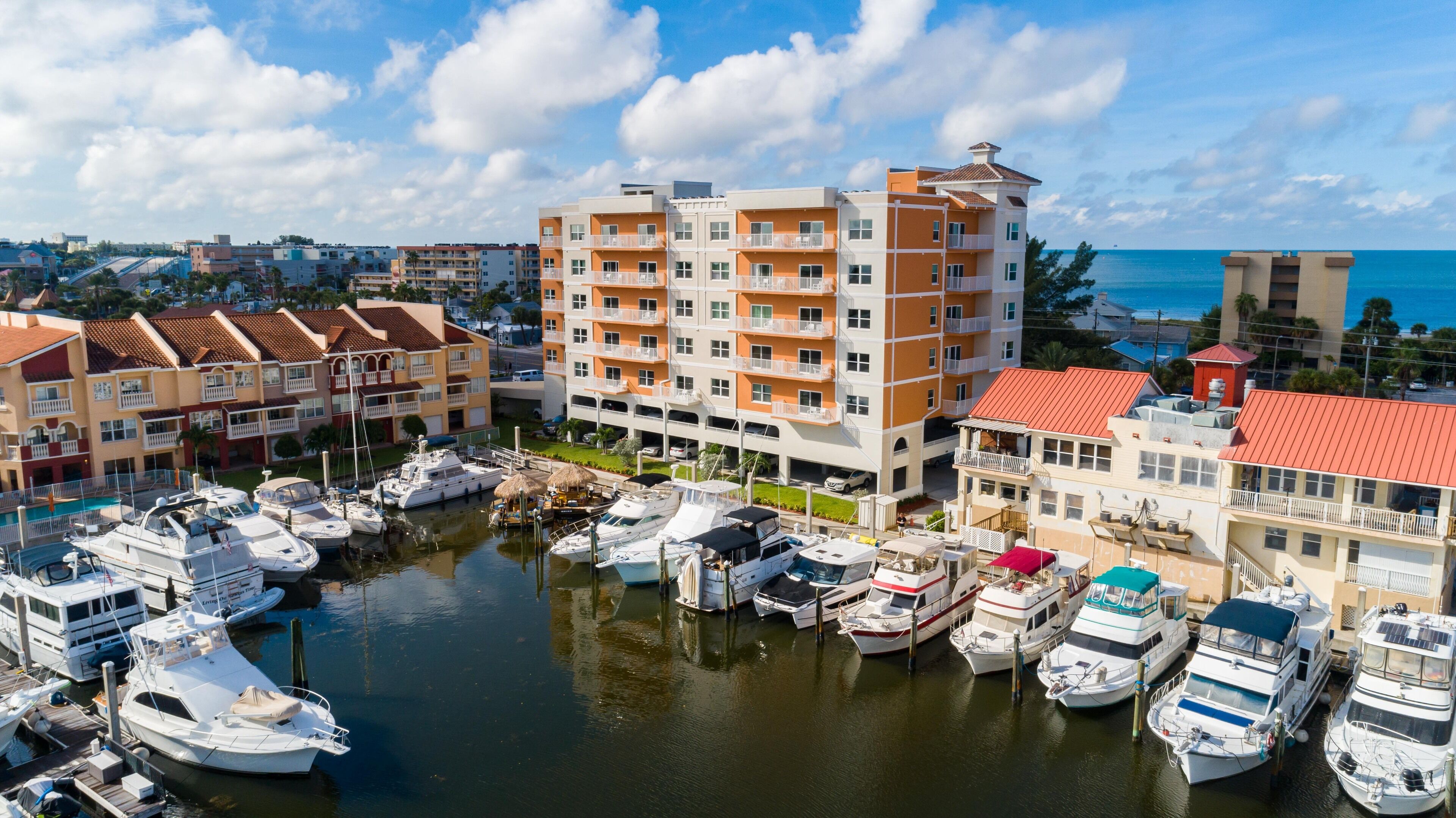 A marina with multiple boats docked, surrounded by multi-story buildings with balconies and a clear view of the sea.