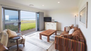 Ein modernes Wohnzimmer mit einem großen Schiebeglasfenster, das auf einen Balkon führt, einem Holztisch, einem gestreiften Sofa und einem Fernseher auf einem weißen Schrank.