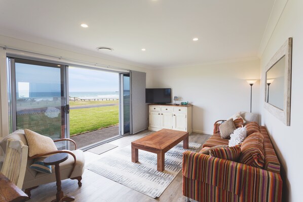 A modern living room with a large sliding glass door leading to a balcony, a wooden coffee table, a striped sofa, and a TV on a white cabinet.