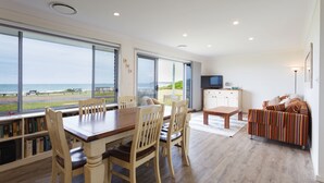 Ein geräumiger Wohnbereich mit großem Esstisch, mehreren Stühlen, einer Couch und einem Fernseher. Große Fenster bieten Blick auf den Strand und einen Patio.