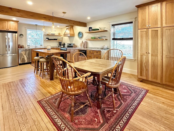 A spacious kitchen with wooden cabinets, a large dining table, and a red patterned rug. - Our Guest House (State College)