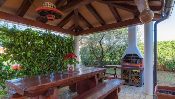 An outdoor patio with a wooden table and benches, a barbecue grill, and a lush green hedge in the background. - Apartment Laura by Interhome (Pula/Galižana)