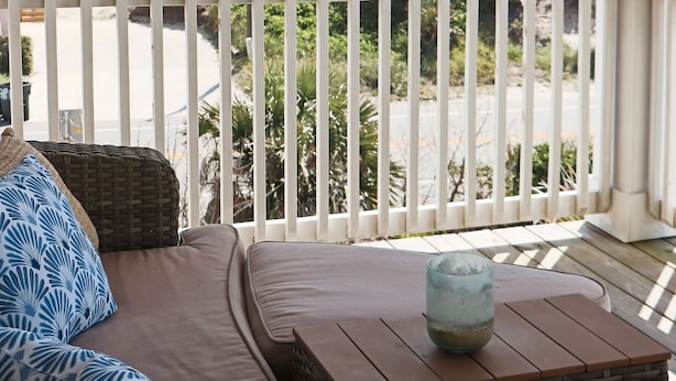 Un balcón con vista al océano, una zona de estar con cojines y una mesa de madera con un vaso.
