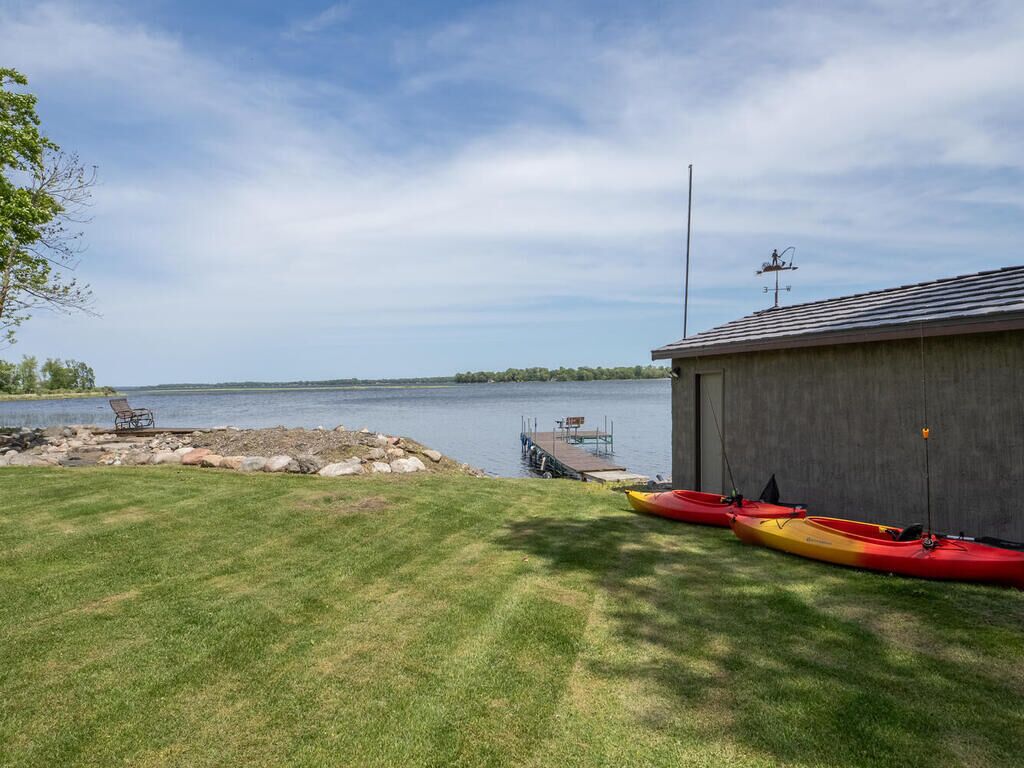 A grassy area with kayaks and a dock extending into a lake, a building with a metal roof, and a clear sky.