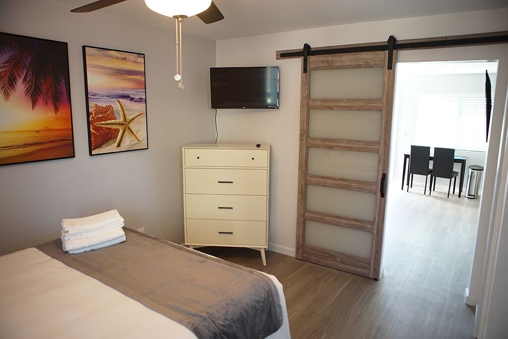 Quarto com cama, cômoda, televisão e porta deslizante para uma varanda.