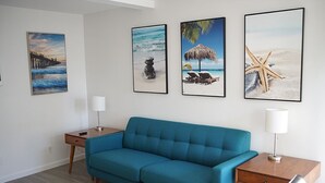 Ein Wohnzimmer mit einem türkisfarbenen Sofa, einem Holztisch, einer Lampe und drei gerahmten Bildern mit Strandmotiv an der Wand.