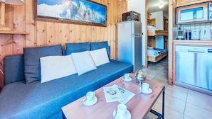 A cozy room with a wooden wall, a blue sofa, a dining table with cups and a spoon, a refrigerator, and a kitchen area with a microwave and kettle.