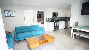 Una sala de estar moderna con un sofá azul, una mesa de centro de madera y una cocina con armarios blancos y un microondas.