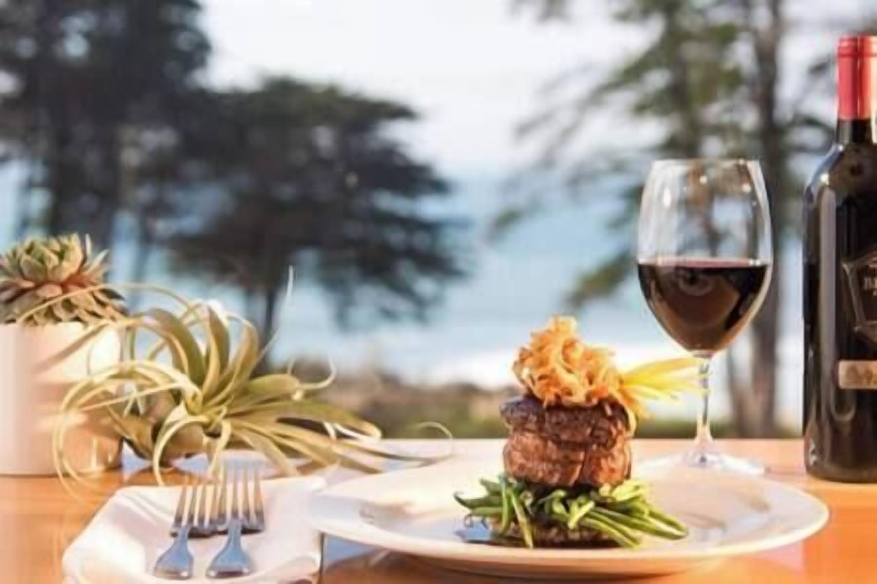 A plate with a burger and green beans, a wine glass, and a bottle of wine on a table with a view of trees and the sky.