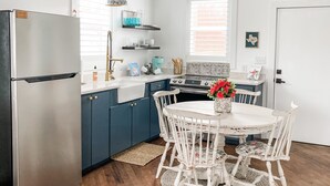A modern kitchen with a stainless steel refrigerator, blue cabinets, a white dining table with white chairs, and a window with blinds.