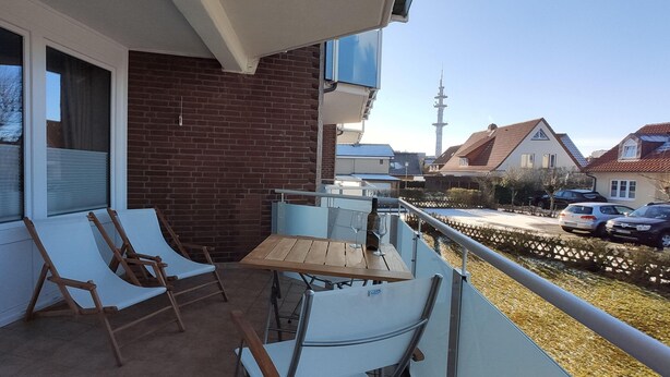 A balcony with two chairs and a table, a brick wall, and a view of a residential area with houses and a tower. - App. Sylter Traumzeit (Sylt)
