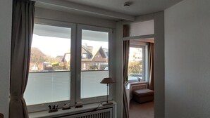 A room with a large window, a radiator, and a small table with a lamp. - App. Sylter Traumzeit (Sylt)