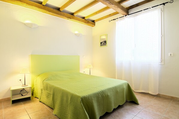 A bedroom with a green bedspread, a nightstand with a lamp, a picture on the wall, and a window with a curtain.