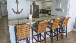 Una cocina con una isla central, cuatro taburetes de mimbre, un refrigerador de acero inoxidable y un letrero que dice "BEACH HOUSE".