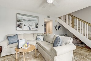 A living room with a beige sectional sofa, a small wooden coffee table, and a framed picture on the wall. A staircase with white railings is visible in the background.
