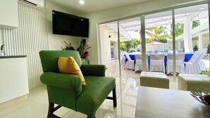 Un fauteuil vert avec un coussin jaune dans un salon moderne équipé d’un téléviseur à écran plat, une porte vitrée donnant sur un espace extérieur avec des tables blanches et des chaises bleues.