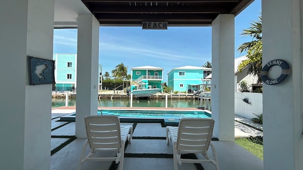 A covered outdoor area with two white chairs, a pool, and turquoise buildings in the background.